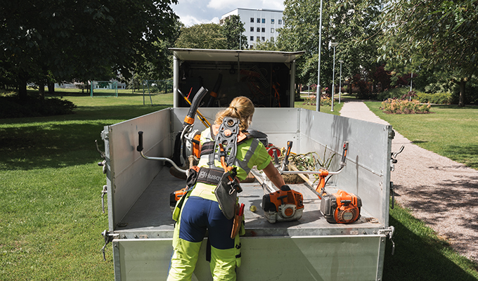 En person iklädd arbetskläder och selar för stora verktyg håller på att lasta ur sin lastbil. Hon är i en stadsnära park och ska arbeta.