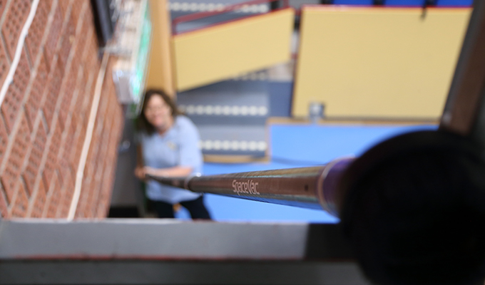 En person använder ett teleskopiskt verktyg för att städa högt upp i en sporthall. Bilden har fokus på verktyget och personen är suddig. 