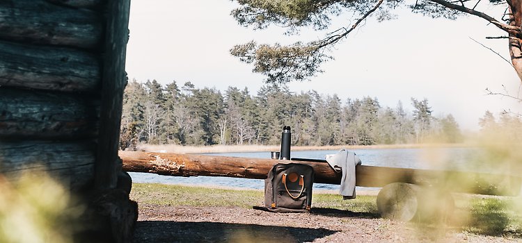 Vindskydd i Ljungnäs, med både vatten och skog i bakgrunden. Vid en bänk gjord av en stam står termos och ryggsäck.