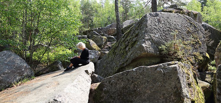 I en skogsmiljö med flera meter höga stenblock sitter en kvinna sitter på ett av de stora stenblocken. 
