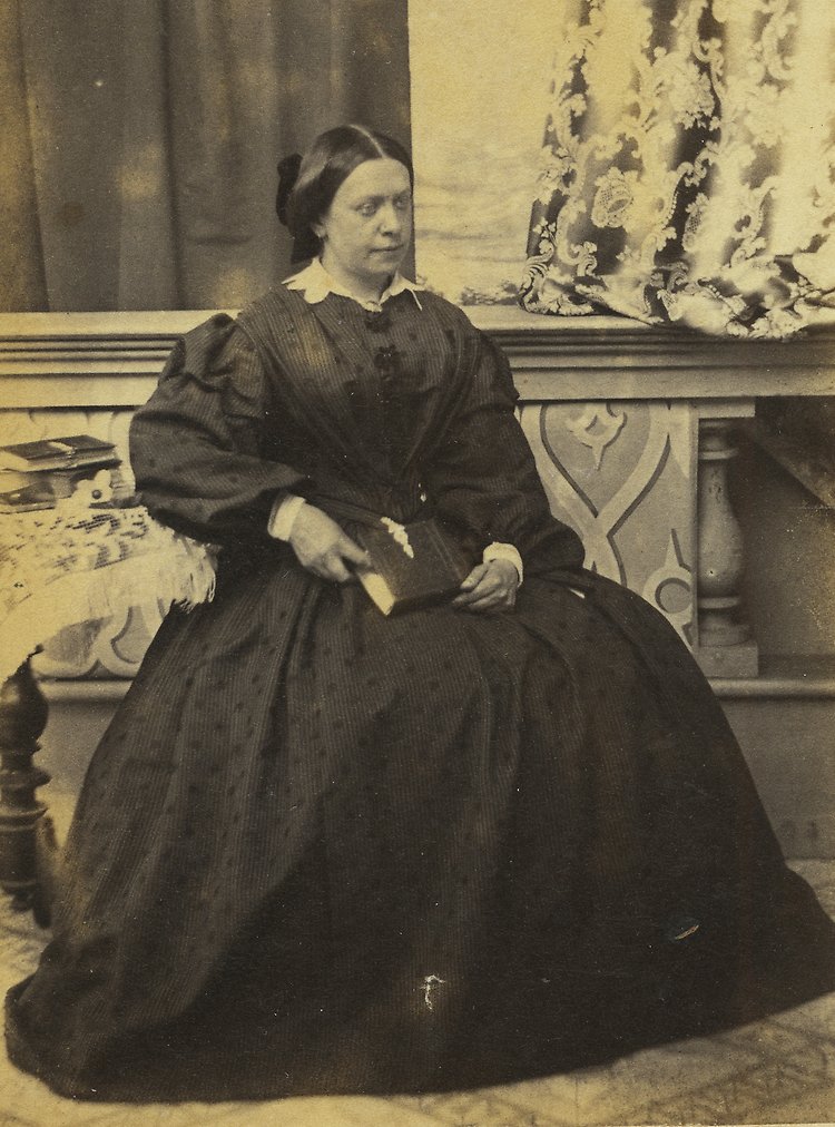 Porträtt på Hermina von Krusenstierna. Hon sitter på en stol i ett dekorerat rum, är iklädd en långklänning och håller i en bok. 