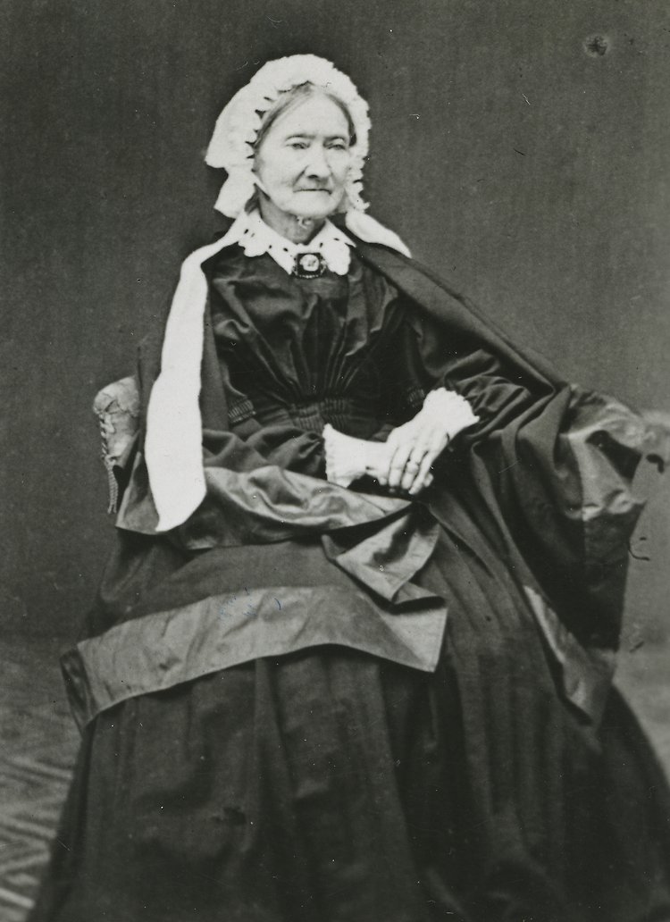 Porträttbild på en äldre kvinna, Herminas mamma. Hon poserar på en stol, iklädd en tidstypisk klänning och har händerna placerade i knät, på varandra.